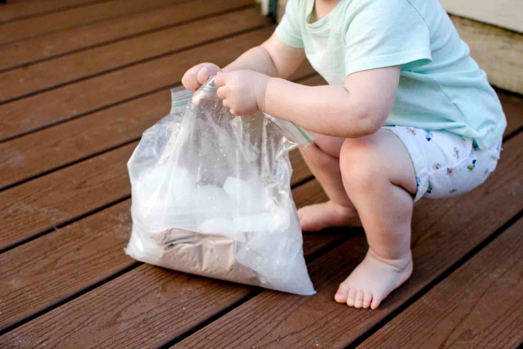 Yeni yürümeye başlayan bir çocuk plastik bir torba tutarak çömeliyor. Çantada dondurma yapmak için onu sallıyor.