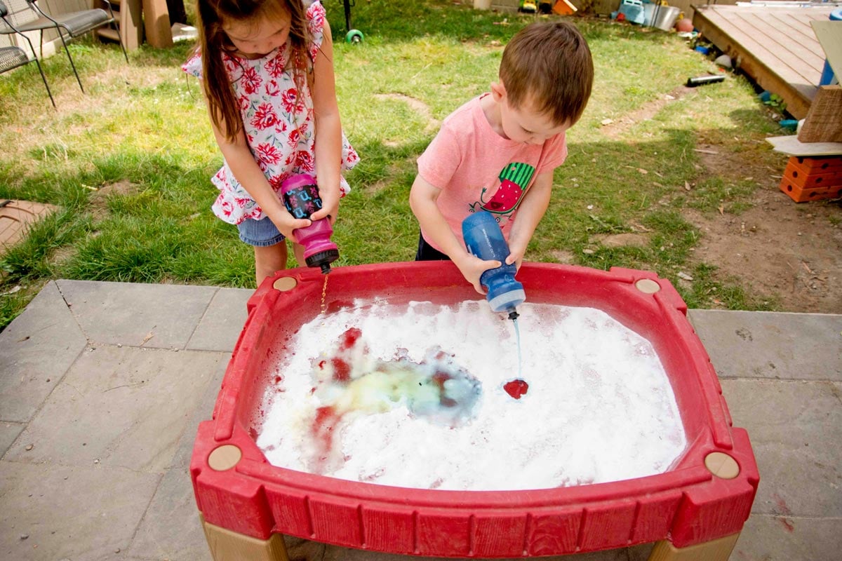 Dışarıda kabartma tozuyla dolu bir su masasının başında renkli sirkeyi sıkmak için su şişeleri kullanan iki çocuğun üstten çekilmiş fotoğrafı.
