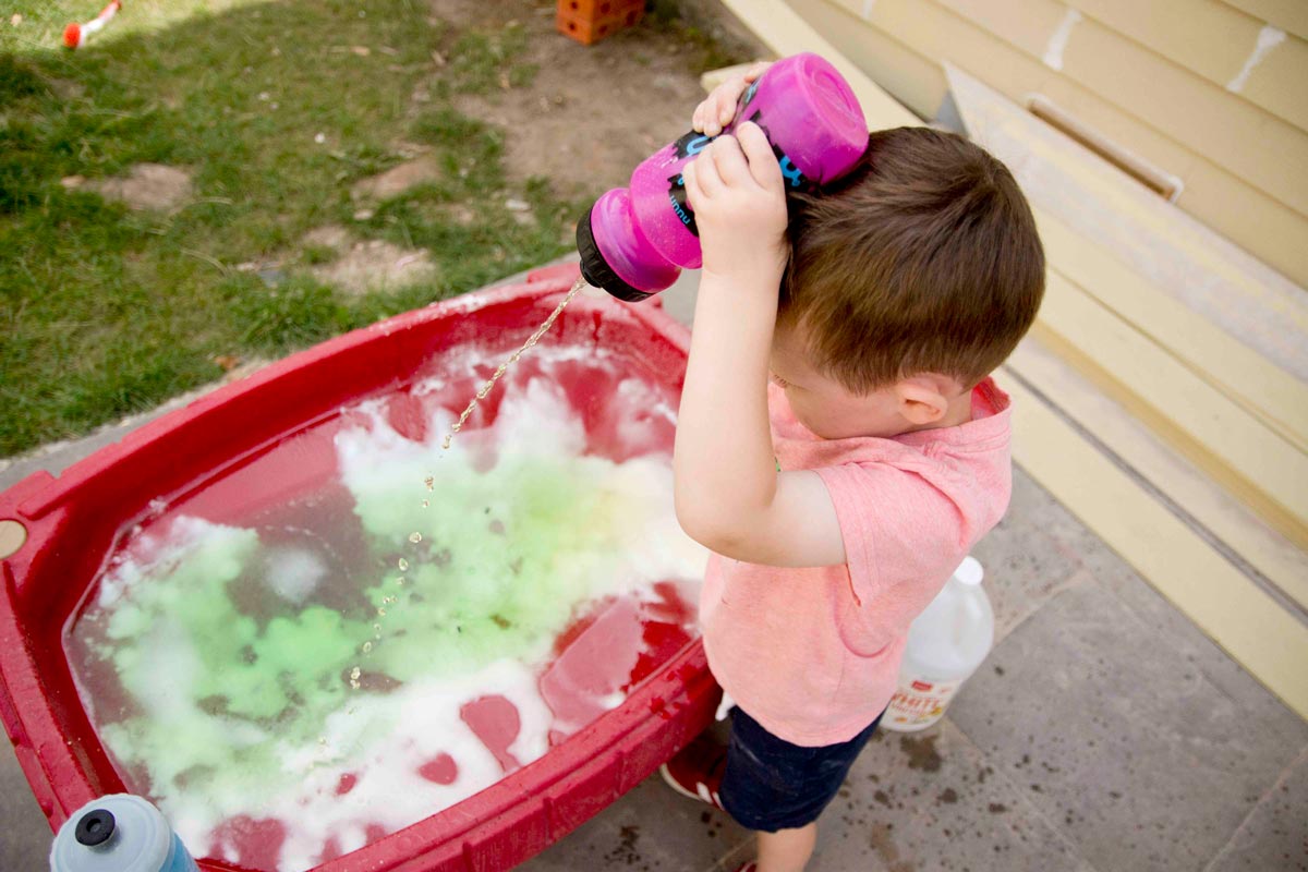 İki yaşında bir çocuk, başının üzerinde bir su şişesi tutuyor ve sirkeyi kabartma tozu dolu kum masasına sıkıyor.